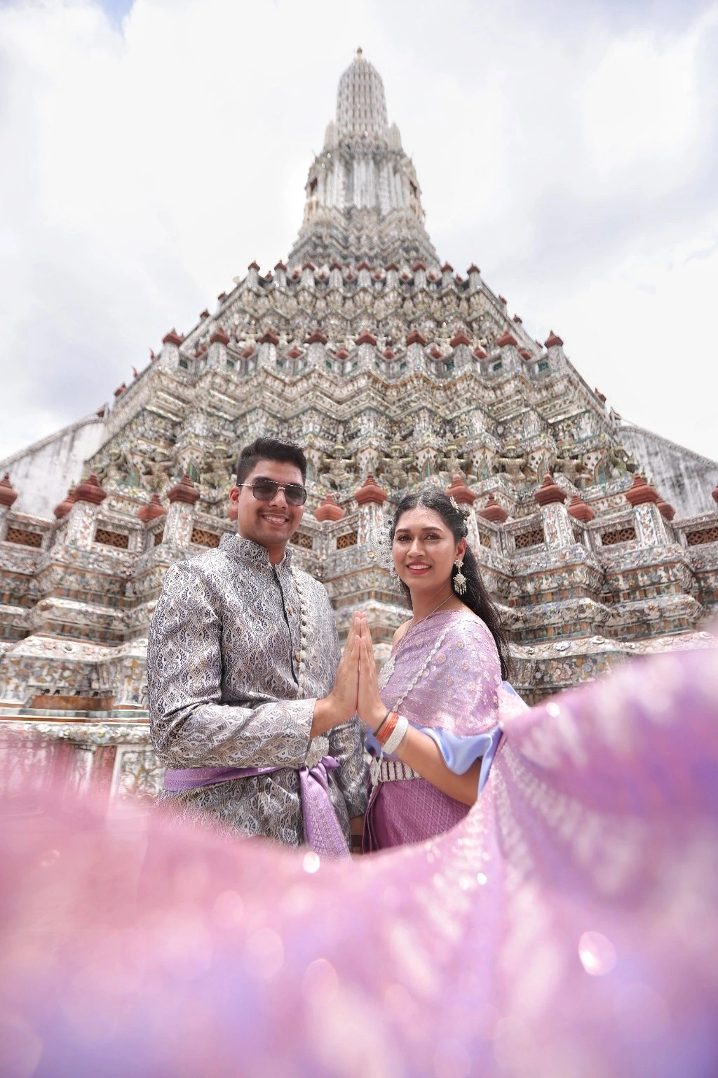 Malika and Jyanesh standing together in front of an ornate temple, framed by a lavender veil